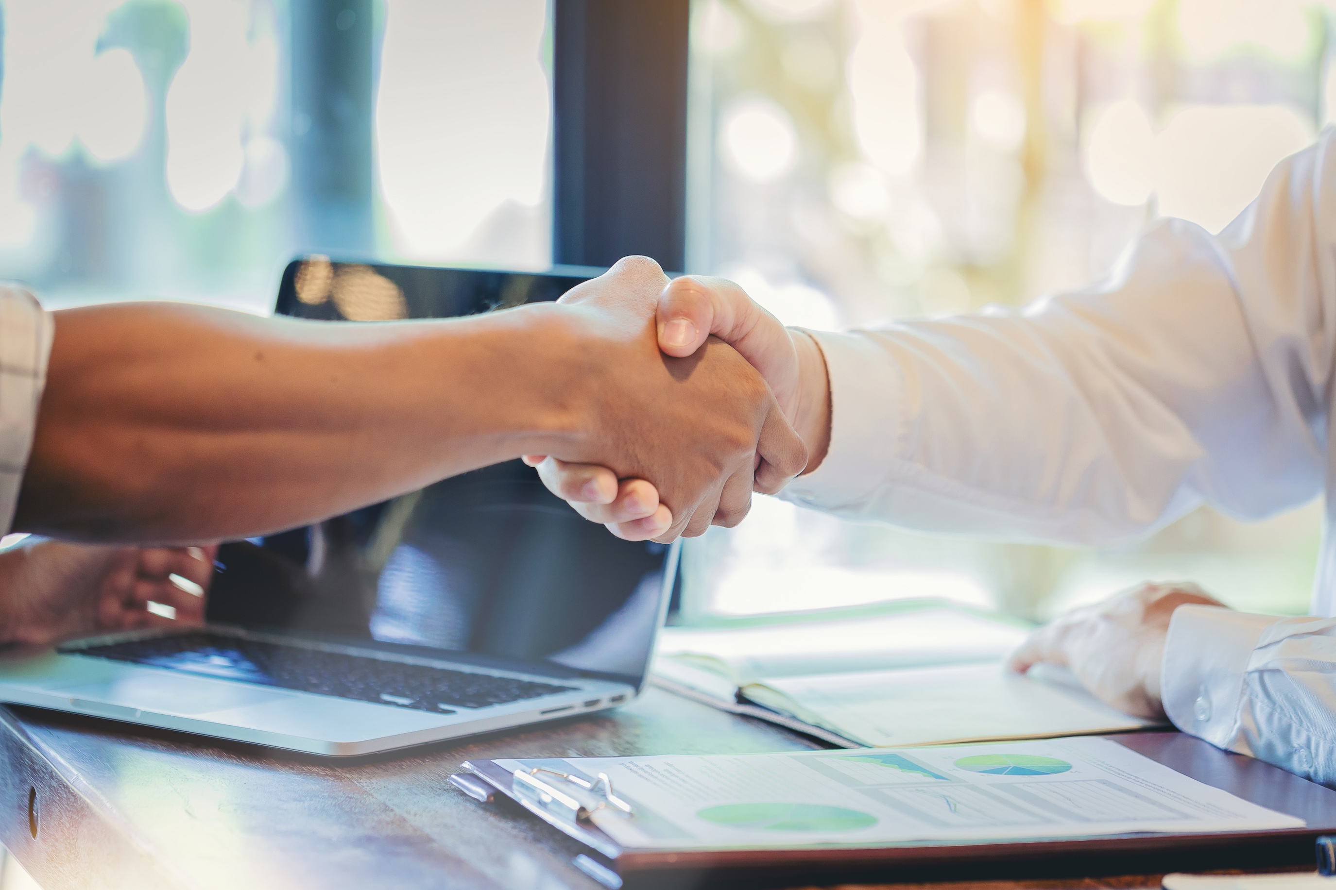 Two individuals in business shirts shaking hands over paperwork on a desk. There is also an open laptop in the background.