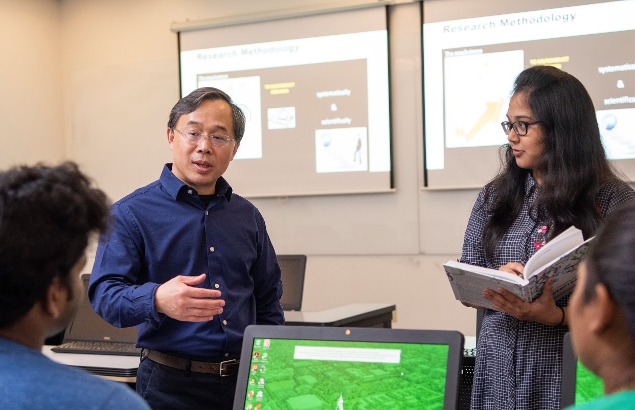 A professor teaching a small group of students with a PowerPoint Presentation in the background.