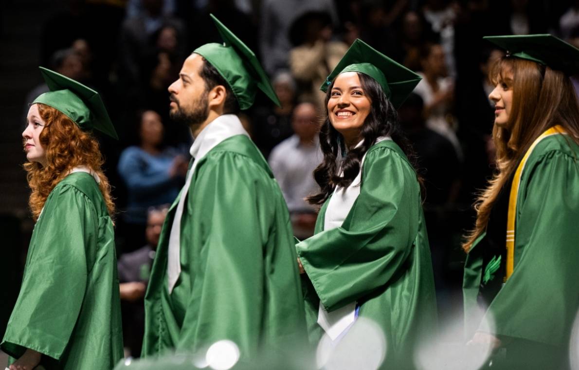 Four UNT graduates in green caps and gowns walking down the aisle at commencement.
