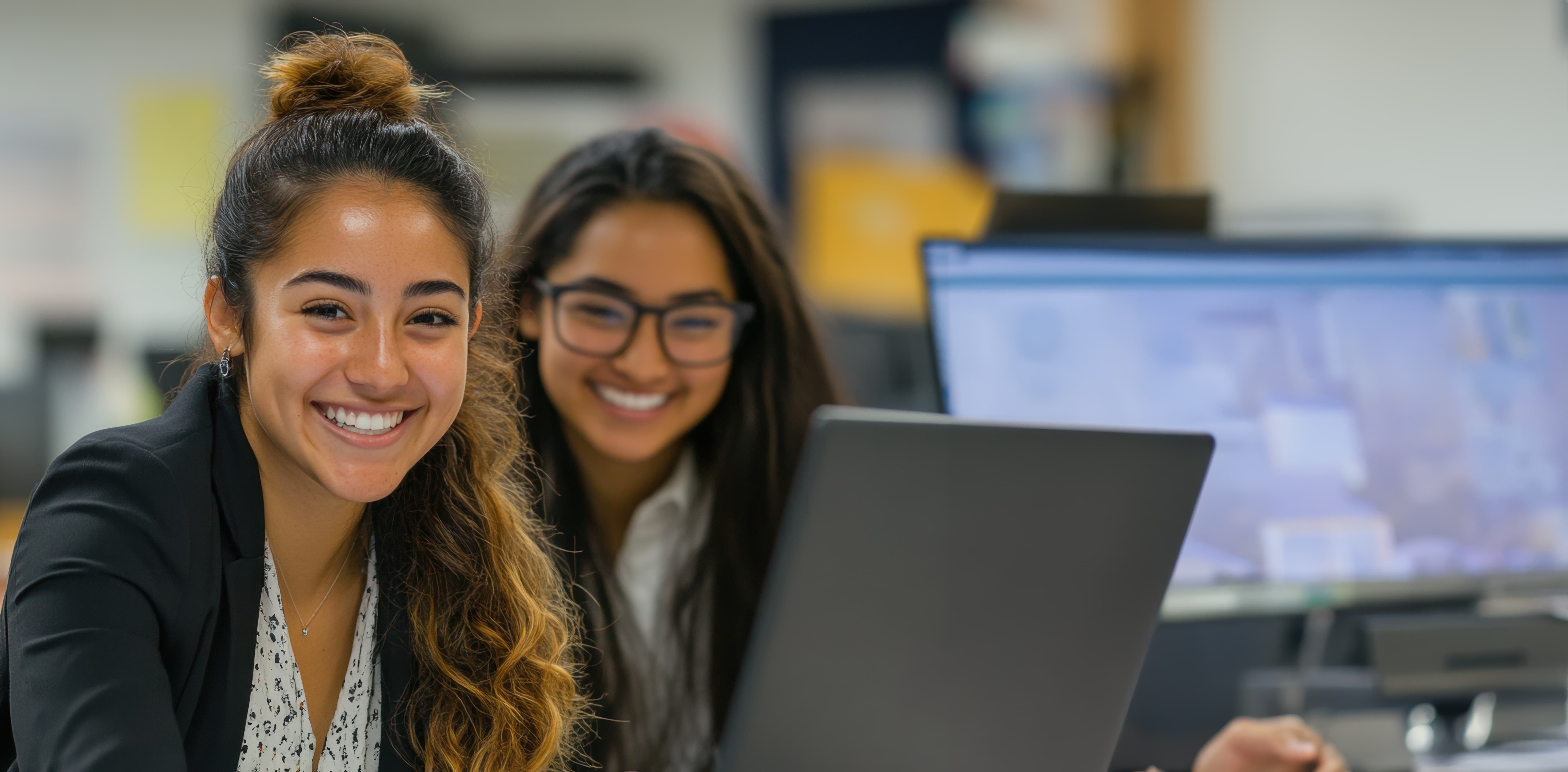 Two female students in professional clothes working on a laptop and other computers while smiling at the camera