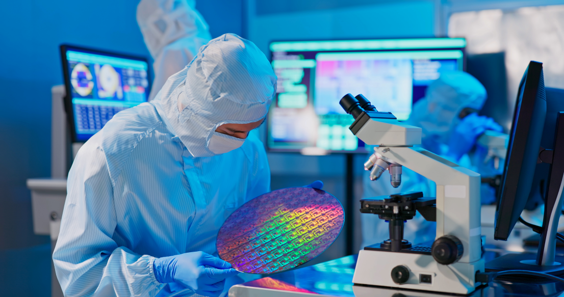 Individual in a white cleanroom lab suit, holding a large circular piece of holographic metal next to a microscope.